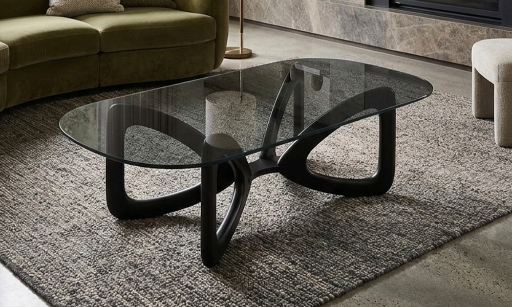 Angelica modern orta sehpa salon yerleşimi; optik ferahlık sağlayan oval şeffaf cam tabla ve heykelsi kavisli mat siyah taşıyıcı iskelet tasarımı.