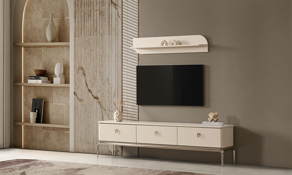 Krem modern TV ünitesi, parlak metal ayakları, üç çekmecesi, duvar rafı ve televizyonla şık salon dekorasyonunda.